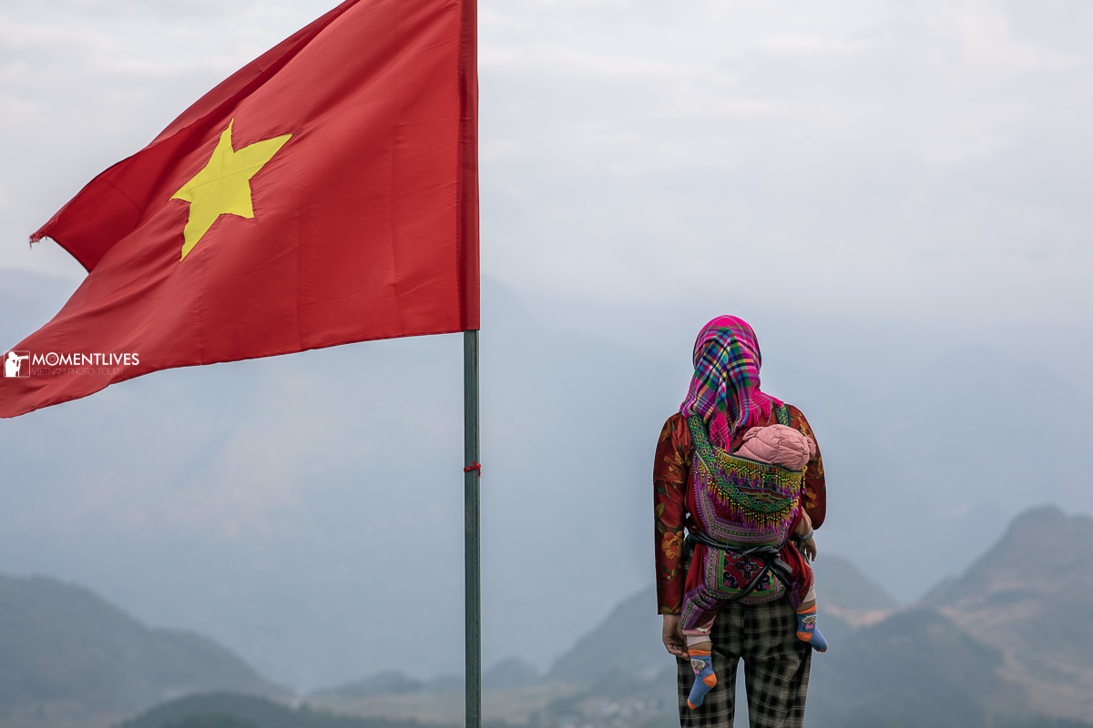 A mother carrying a child standing on a mountain ridge of Ha Giang with the Vietnamese flag next to them
