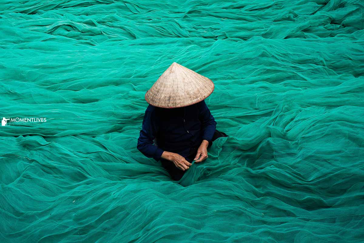 A lady wearing a conical hat, sitting on a surface of fishing net to mend