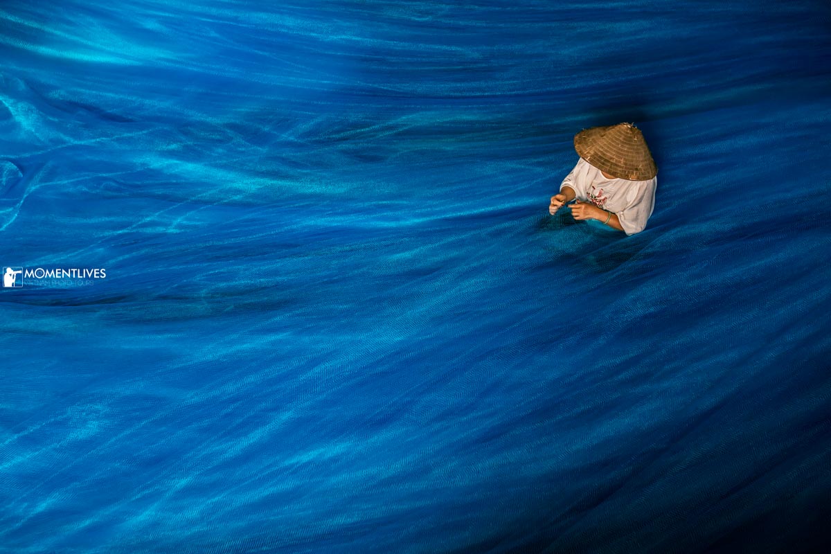 A Vietnamese woman wearing a conical hat sitting and mending a fishing net